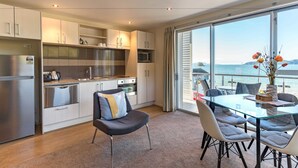 A modern kitchen with stainless steel appliances, a dining area with a glass table and chairs, and a large window overlooking a body of water. - Edgewater Palms Apartments (Paihia)