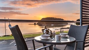 A balcony with a table set for tea, overlooking a calm body of water and a distant island during sunset. - Edgewater Palms Apartments (Paihia)