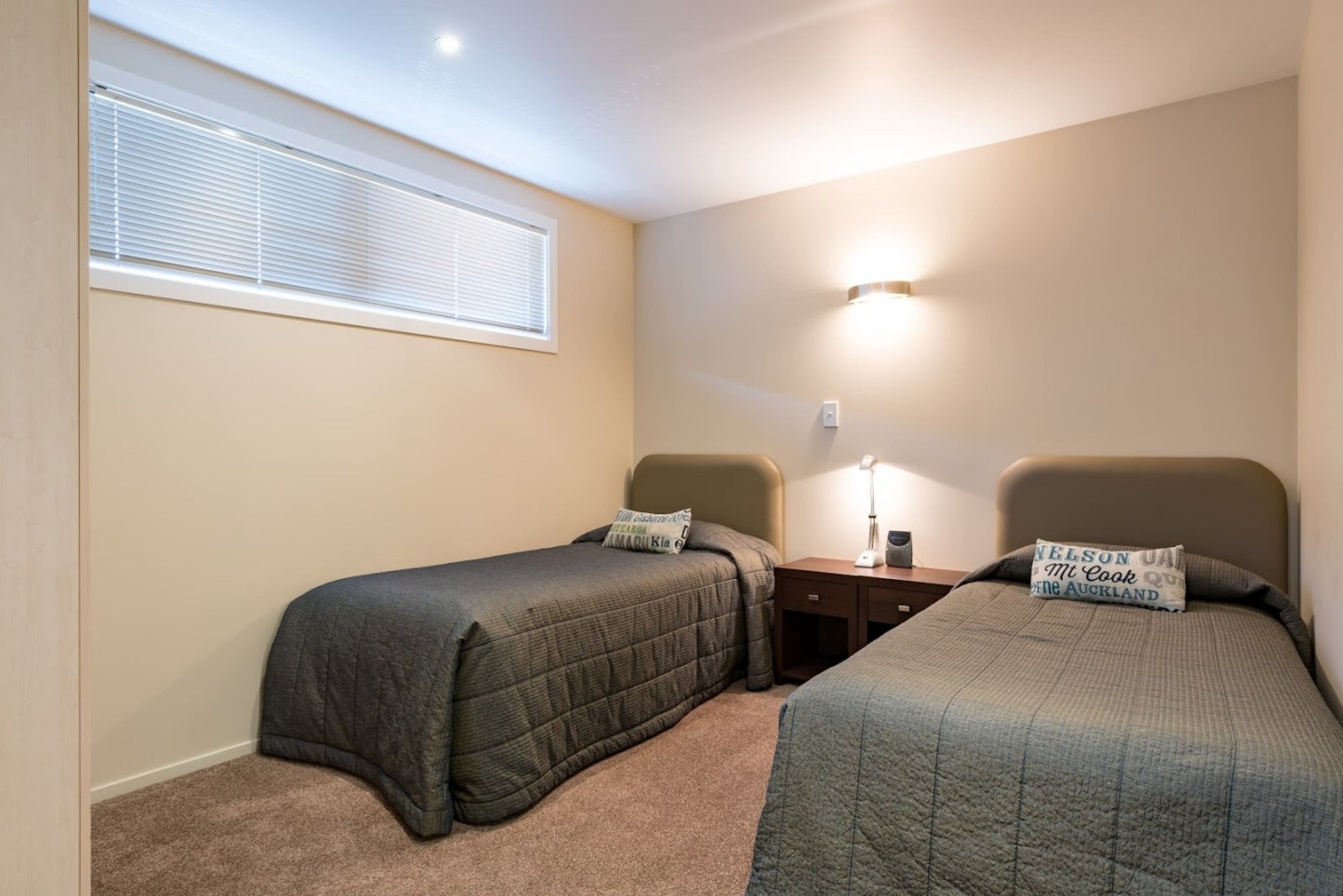 A bedroom with two single beds, a nightstand, a lamp, and a window with blinds.