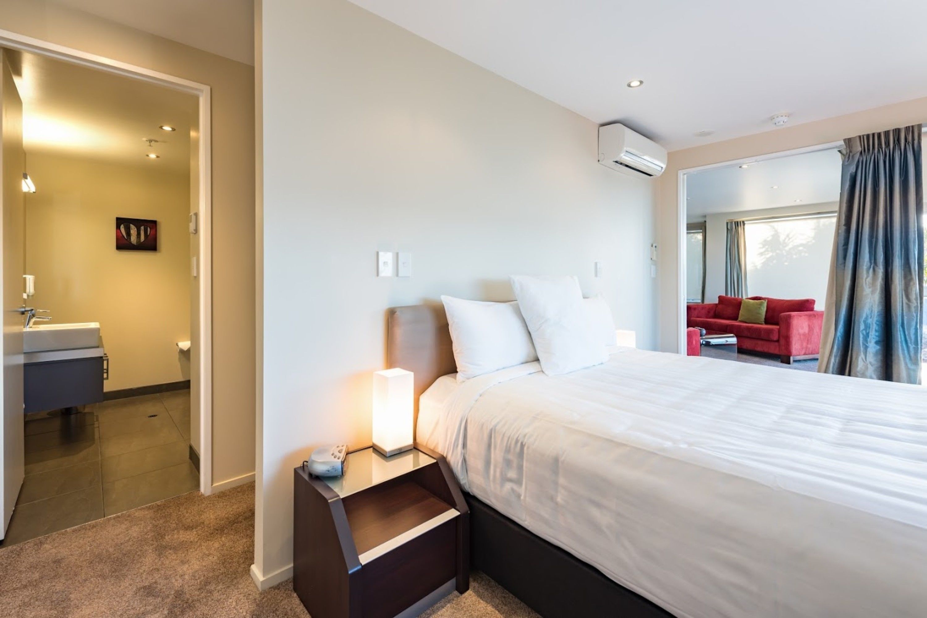 A hotel room with a bed, bedside table, and a red couch in the background.