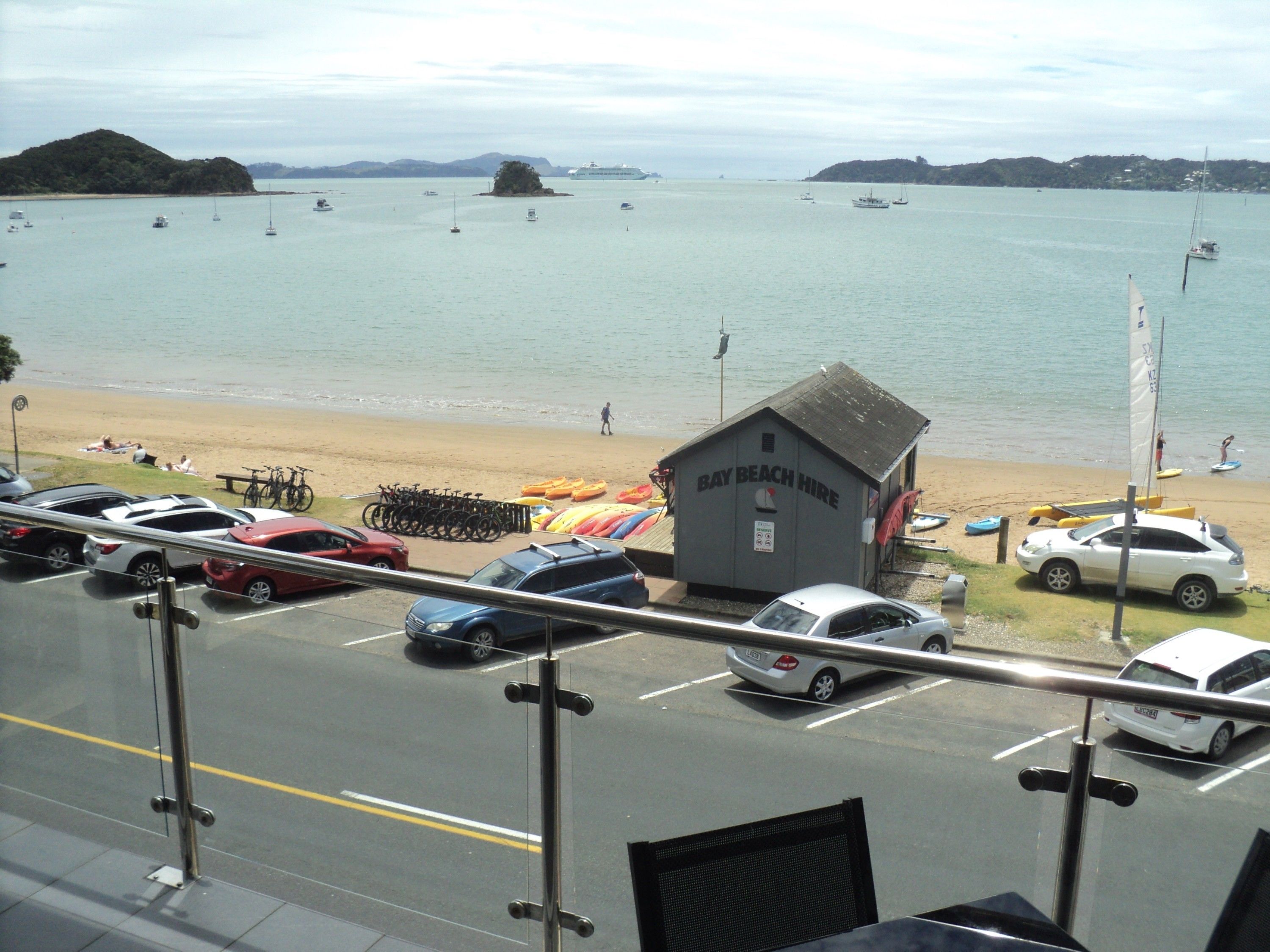 A view of a beach with a small building labeled "Bay Beach Hire" and several parked cars along the roadside.