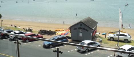 A view of a beach with a small building labeled "Bay Beach Hire" and several parked cars along the roadside.