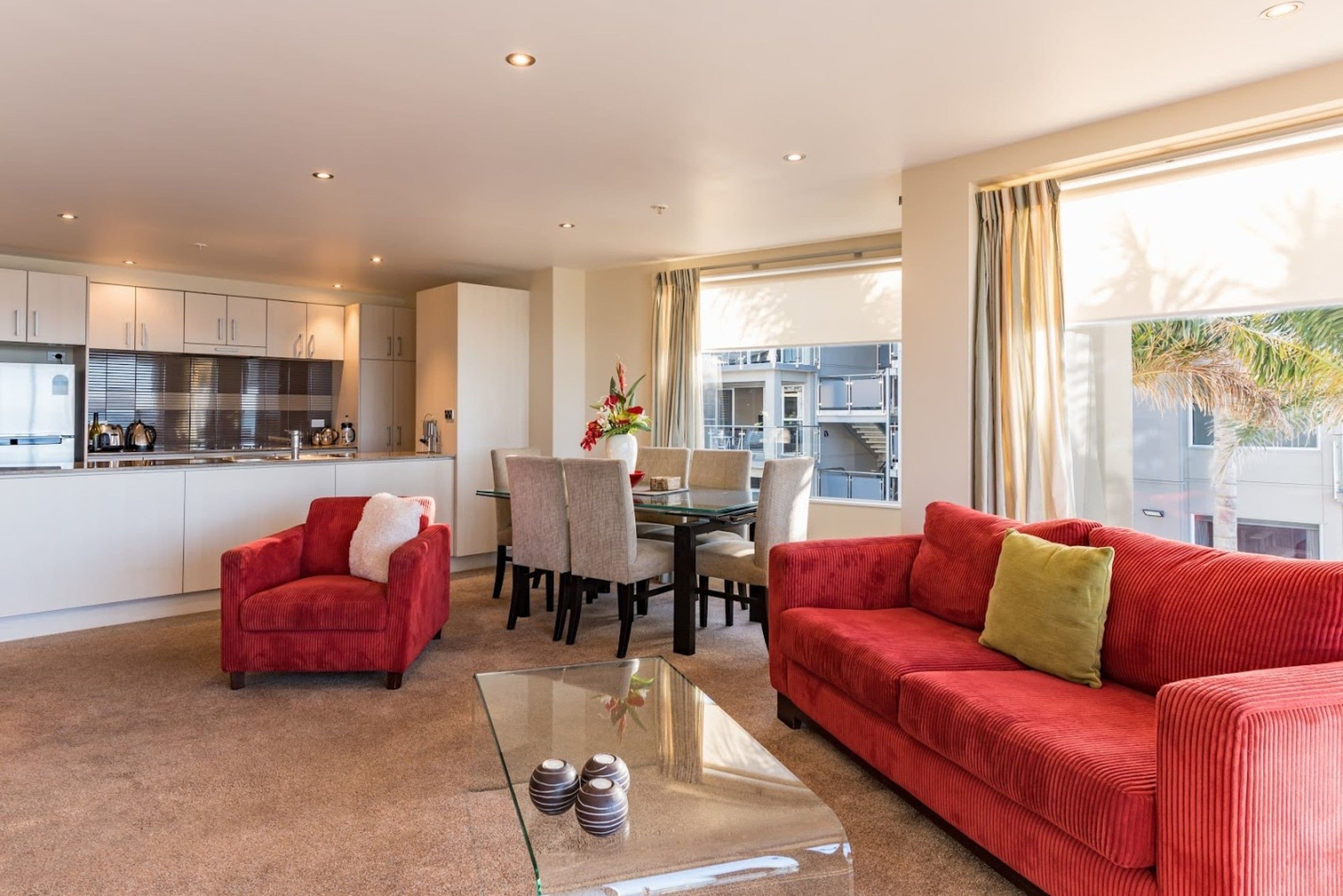 A modern living room with a glass coffee table, red sofas, a dining area with a glass table and chairs, and a kitchen with stainless steel appliances.