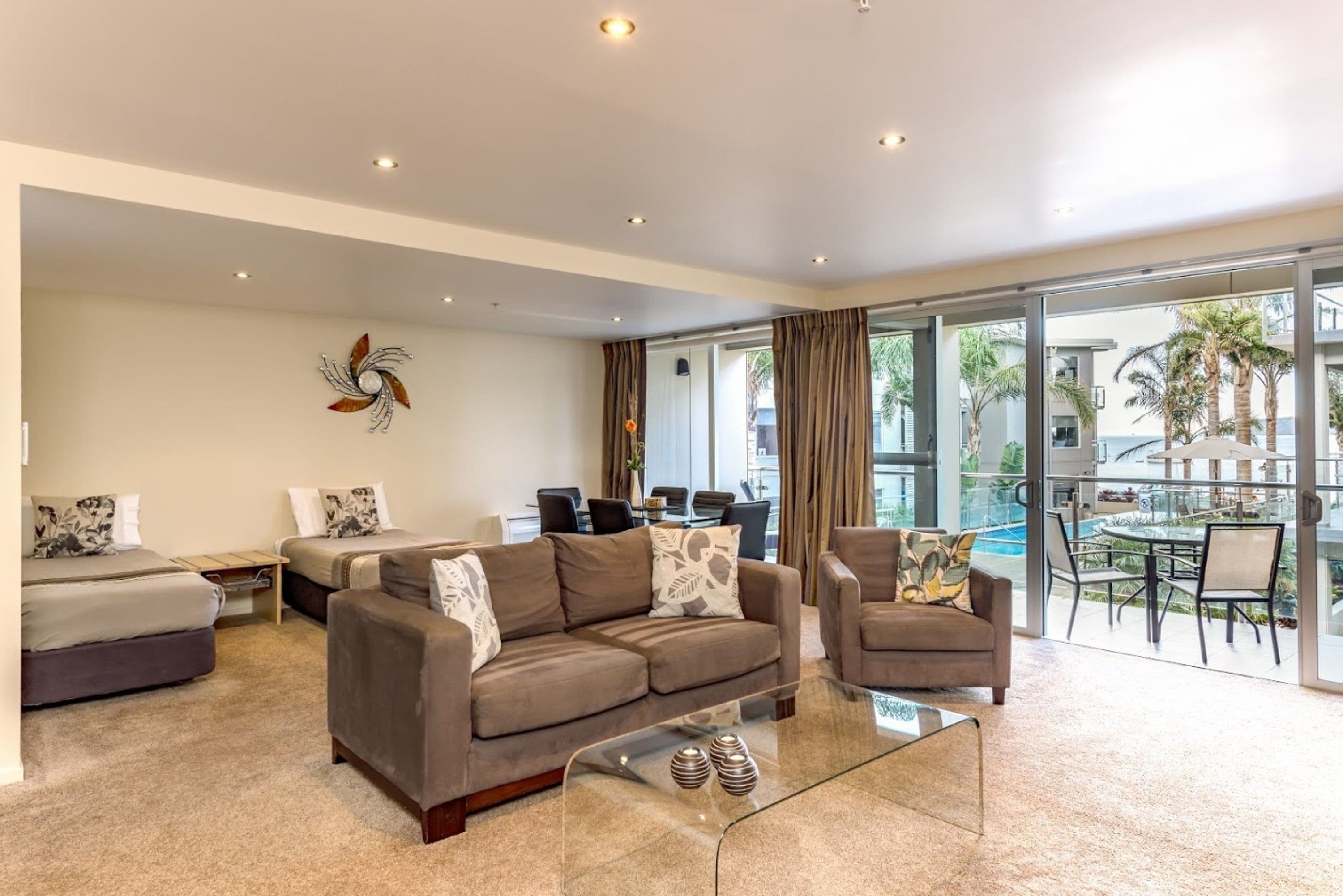 Uma sala de estar moderna com sofá, poltronas e uma mesa de centro de vidro. Há uma área de jantar com mesa e cadeiras, e uma vista para uma piscina e palmeiras através de grandes janelas.