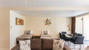 A modern hotel room with a brown sofa, two beds, a dining table with chairs, and a decorative wall piece.
