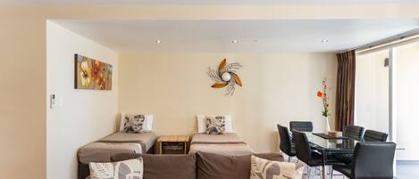 Une chambre d’hôtel moderne avec un canapé marron, deux lits, une table à manger avec des chaises et un élément décoratif mural.