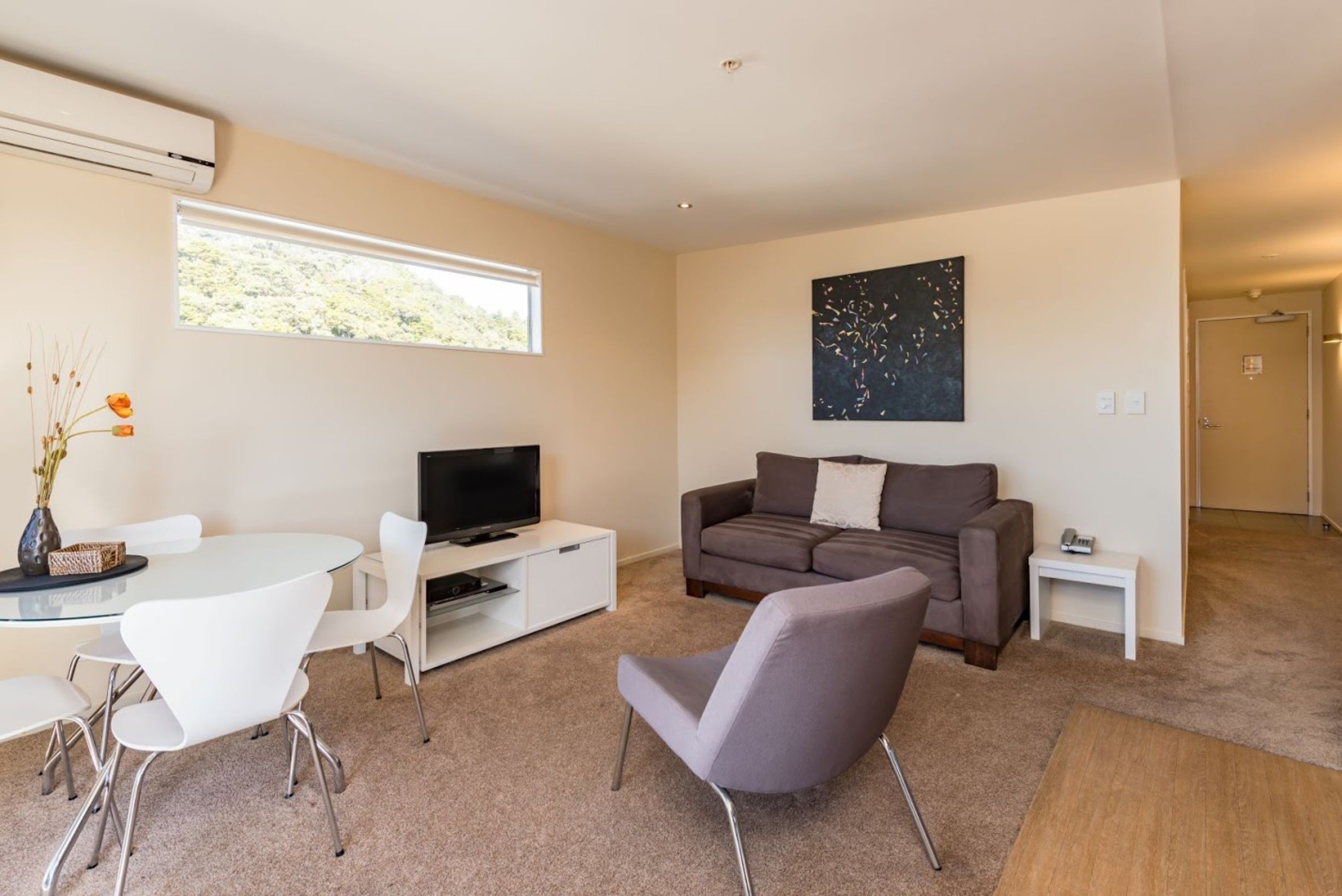 A modern living room with a flat-screen TV, a gray sofa, white chairs, and a glass dining table.