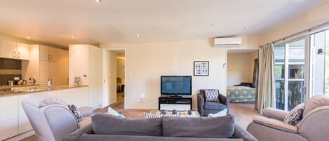 A modern living room with a grey sofa set, a flat-screen TV, and a dining area with a kitchen in the background.
