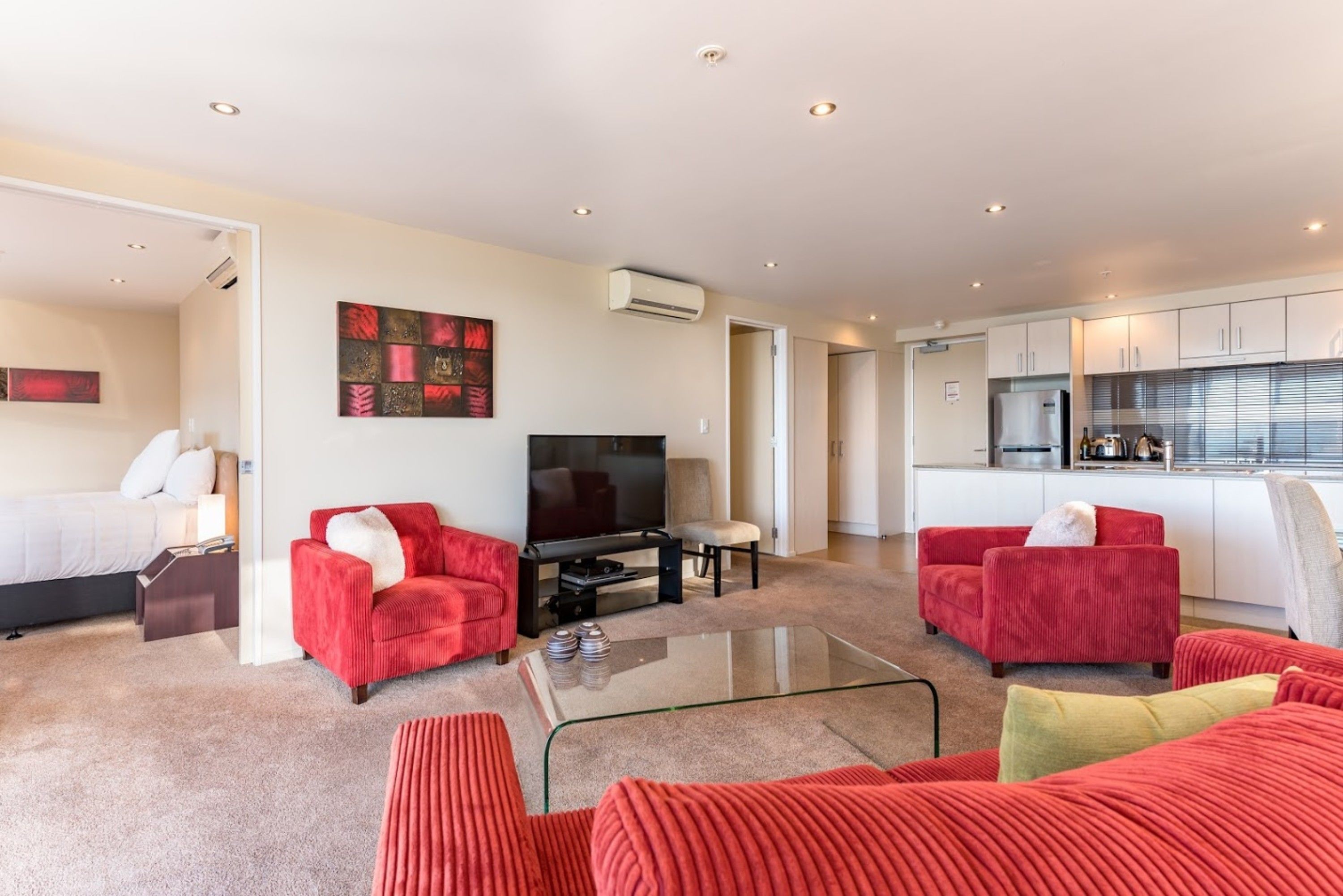 Una sala de estar moderna con sofás tapizados en rojo, una mesa de centro de vidrio, un televisor de pantalla plana y una cocina con gabinetes blancos y un refrigerador.