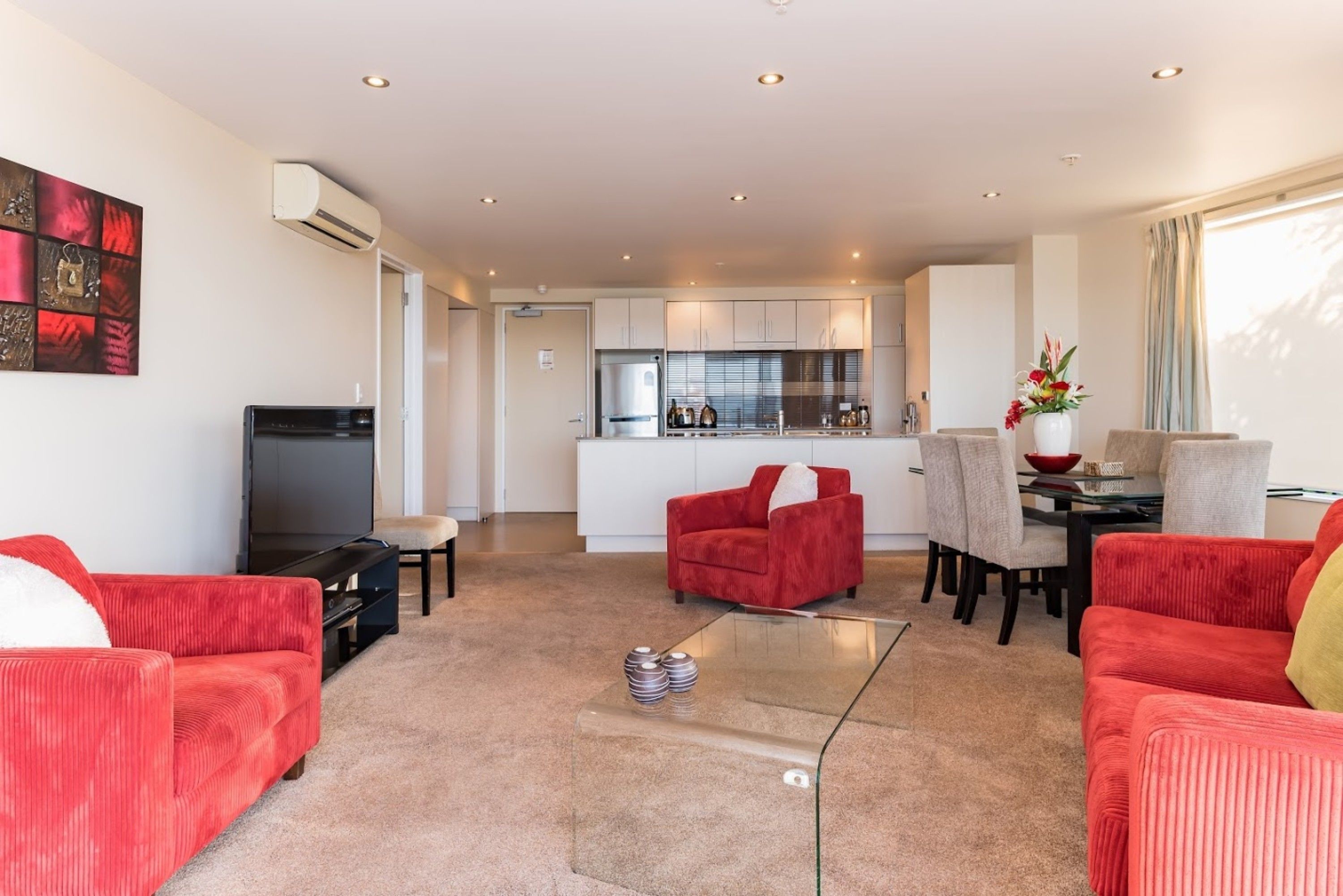 Una sala de estar moderna con sofás rojos, una mesa de centro de vidrio y una cocina al fondo.