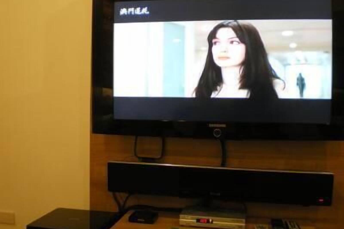 Un televisor montado en la pared, con un control remoto y algunos libros sobre un soporte de madera debajo.