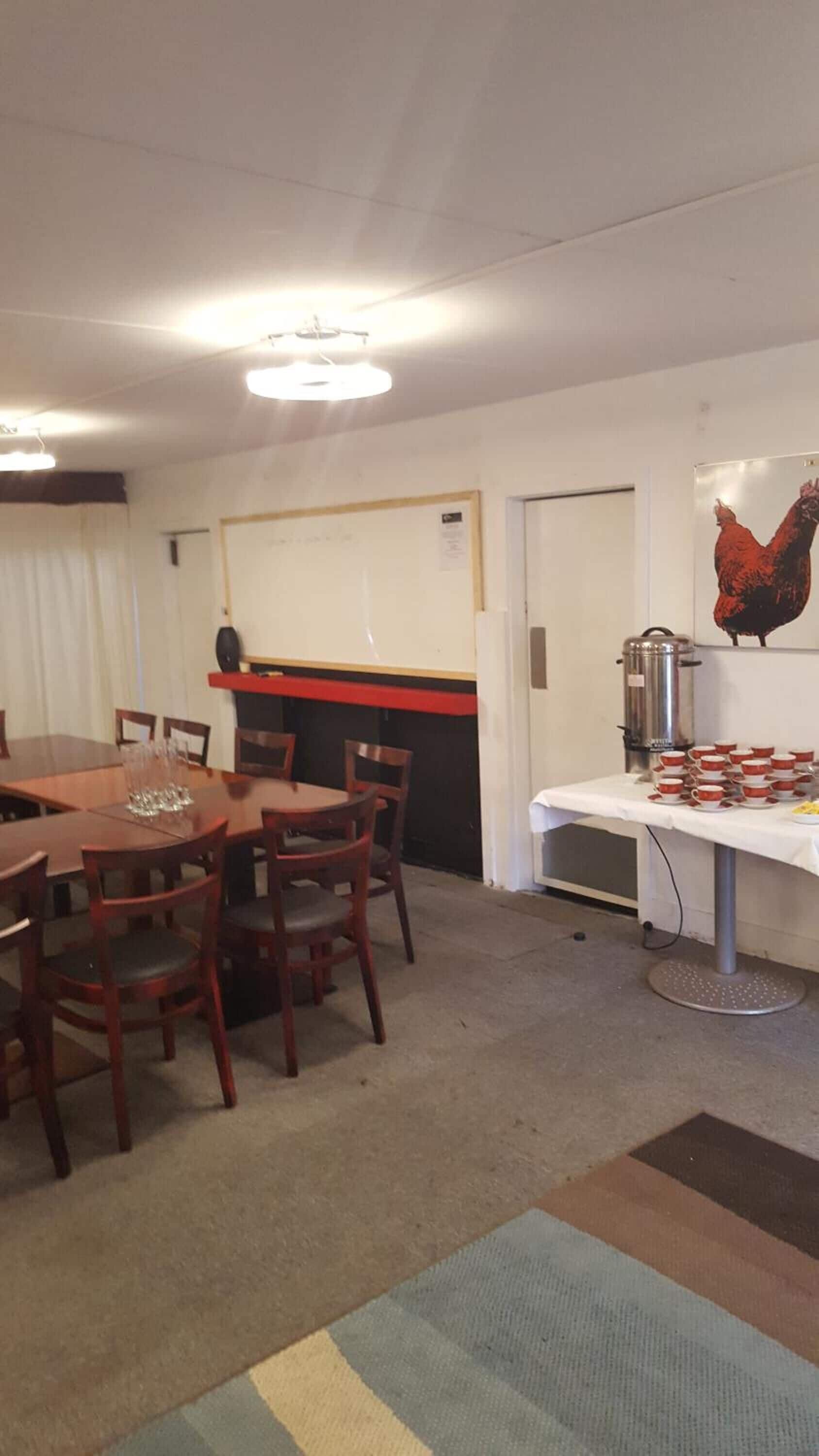A dining area with a long table and chairs, a buffet table with cups and a coffee pot, and a wall with a rooster painting.
