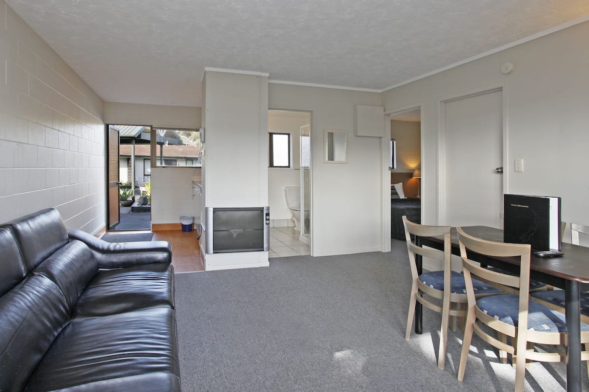 A modern living room with a black leather sofa, a wooden dining table with chairs, and a small kitchen area with a microwave.