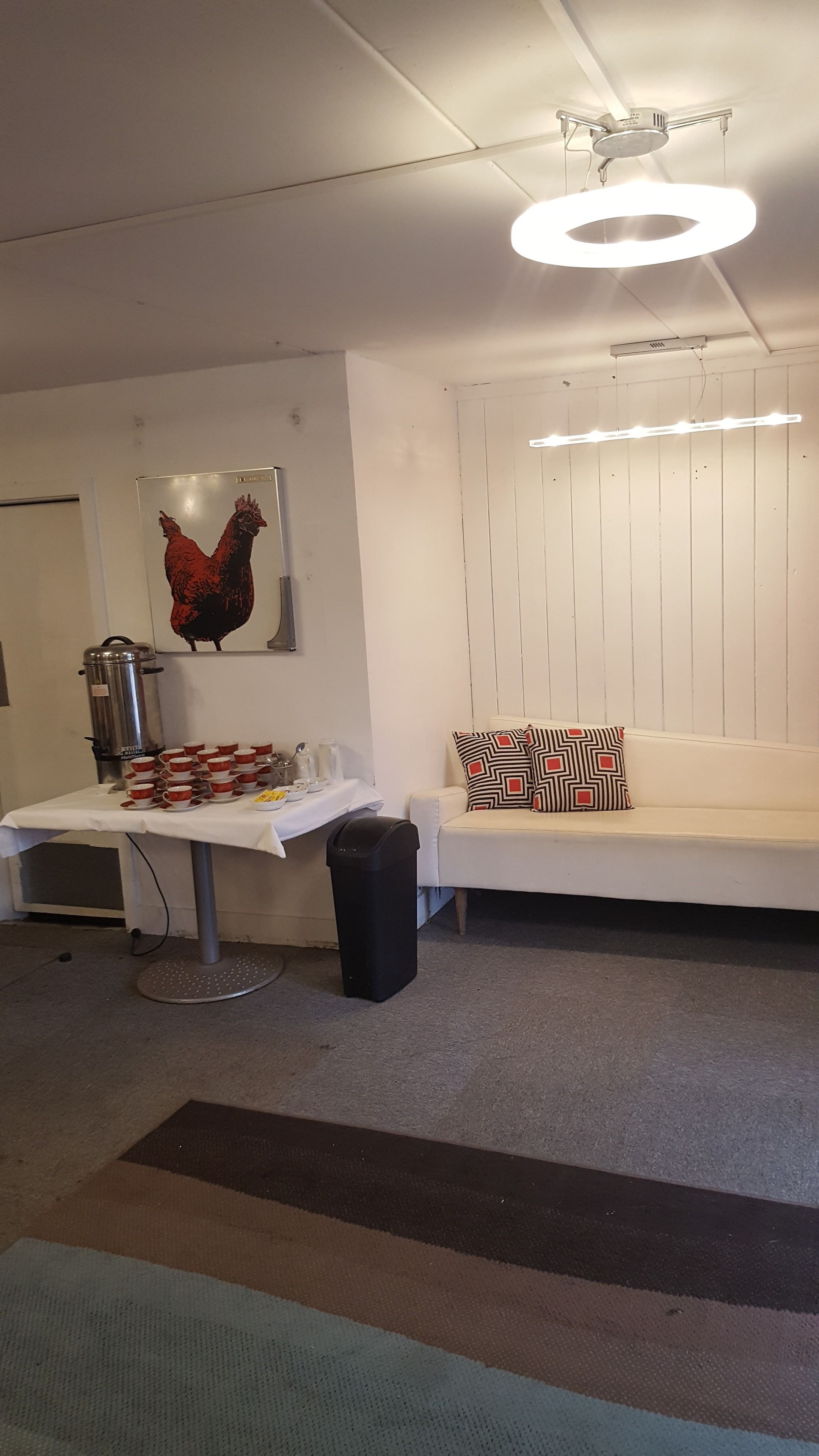 Una habitación con pared blanca, ventilador de techo, una mesa con tazas y una cafetera, un cesto de basura negro y un sofá con cojines estampados.