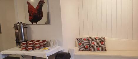 Una habitación con pared blanca, ventilador de techo, una mesa con tazas y una cafetera, un cesto de basura negro y un sofá con cojines estampados.