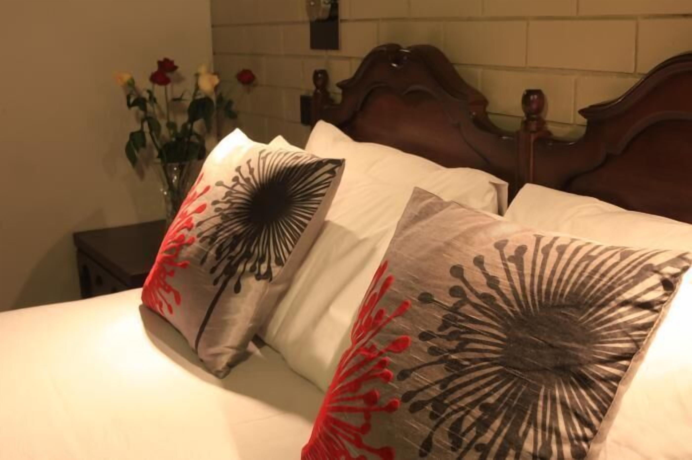A bed with two pillows featuring a black and red floral design, a wooden headboard, and a vase with flowers on a bedside table.