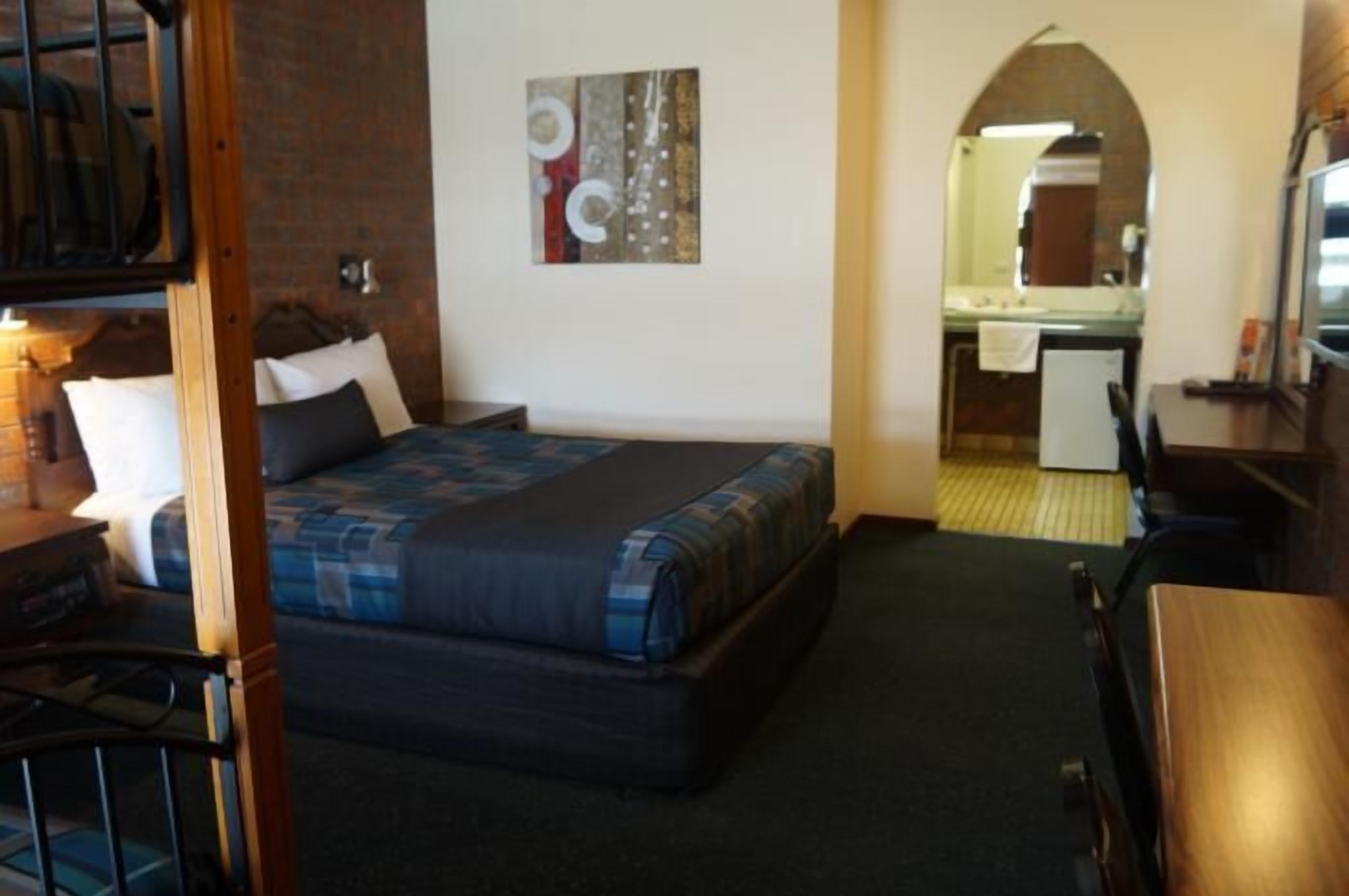 A hotel room with a bed, a desk, a chair, a small table, a mirror, and a bathroom visible through an archway.