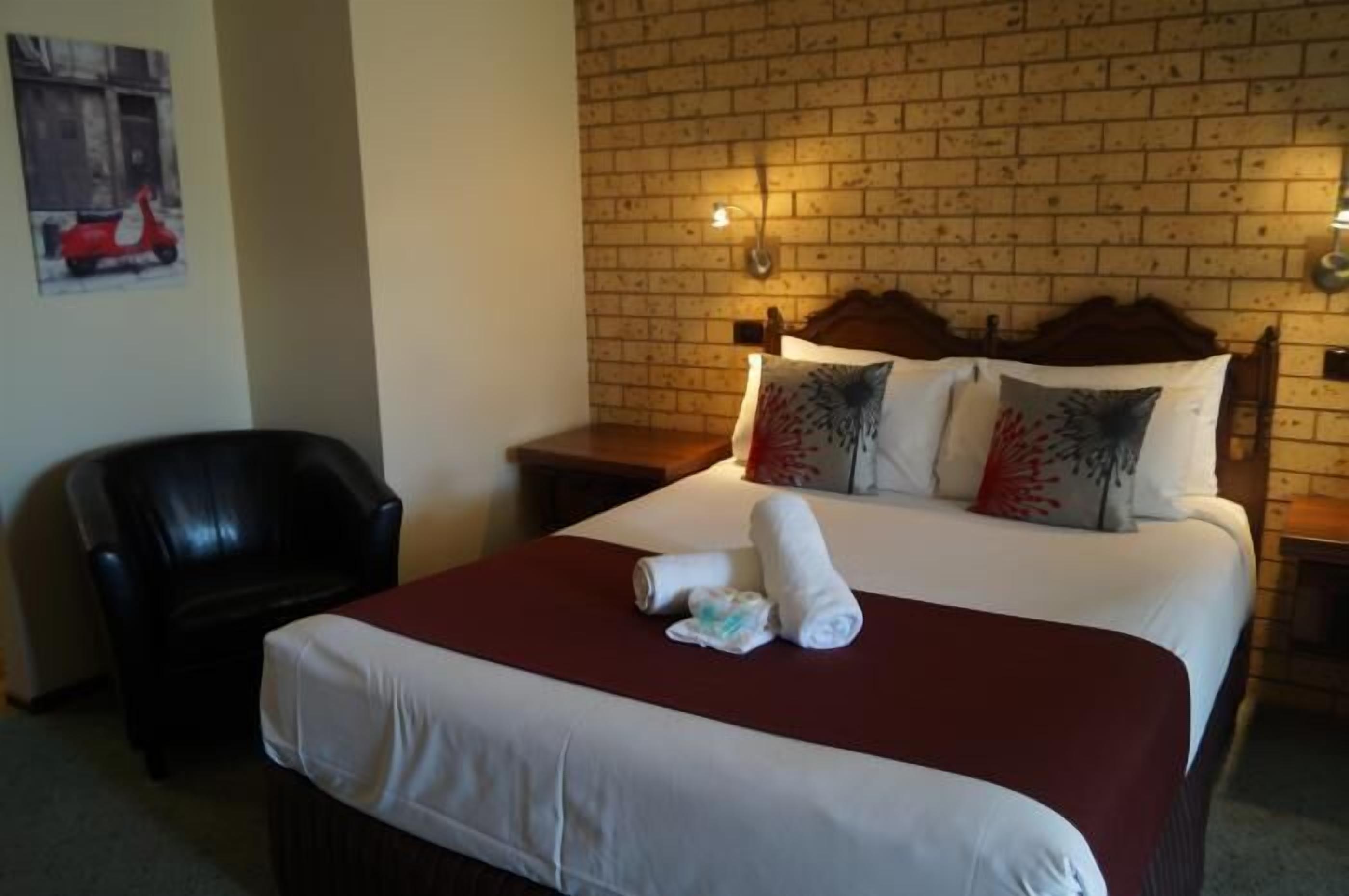 A hotel room with a brick wall, a bed with white and red bedding, a black leather chair, and two bedside tables.
