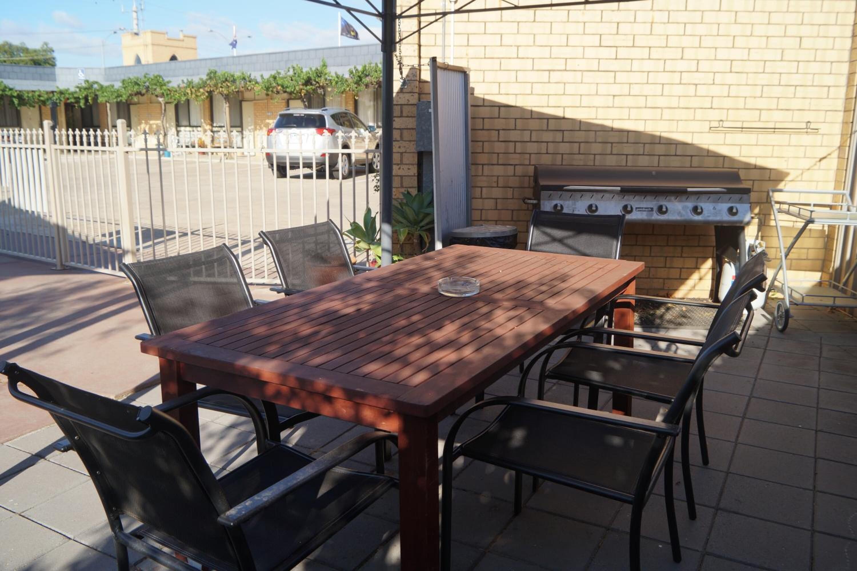 Un patio al aire libre con una mesa de madera y sillas de metal, una parrilla y un muro de ladrillo al fondo.