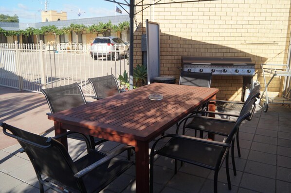 An outdoor patio with a wooden table and metal chairs, a barbecue grill, and a brick wall in the background.