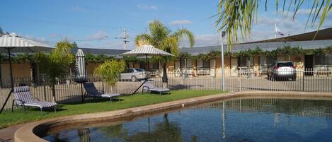 A swimming pool with lounge chairs and umbrellas, surrounded by a fence and greenery.