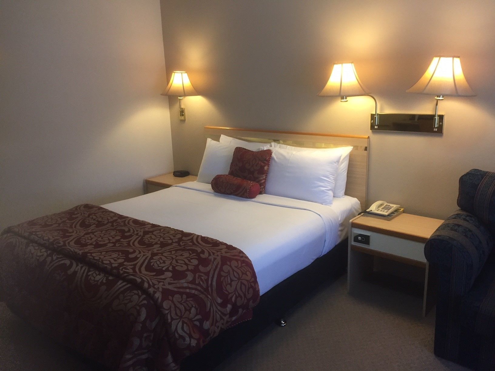 Quarto de hotel com cama, duas luminárias de cabeceira, telefone e uma cadeira.
