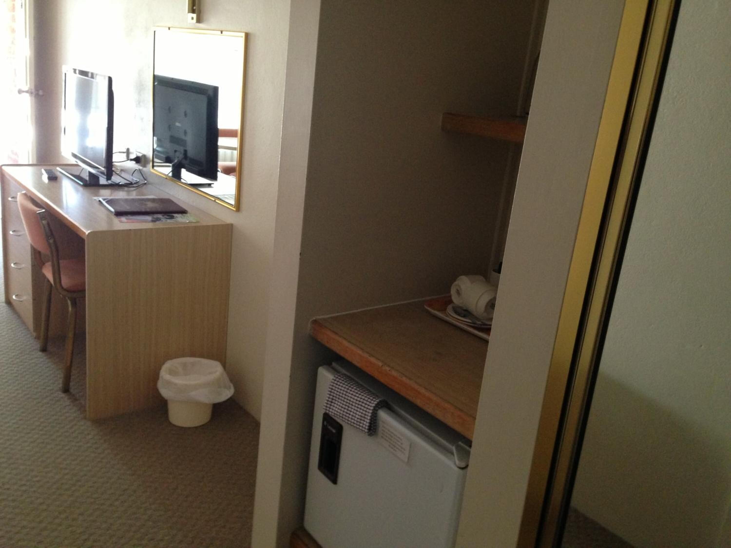 Habitación de hotel pequeña con un escritorio de madera, dos monitores, un cesto de basura, un refrigerador compacto y una sola silla.