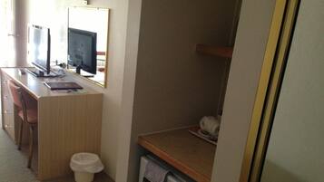 Habitación de hotel pequeña con un escritorio de madera, dos monitores, un cesto de basura, un refrigerador compacto y una sola silla.