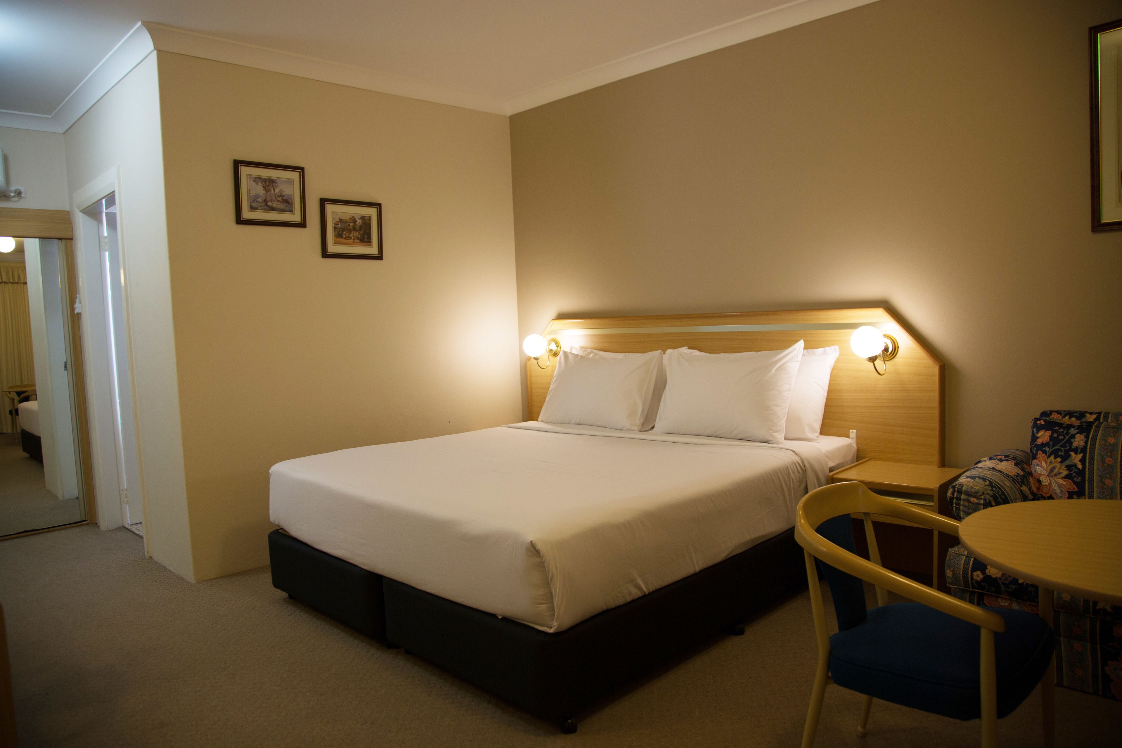 Habitación de hotel con cama, una silla, una mesa y dos cuadros en la pared.