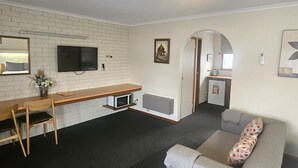 Un soggiorno moderno con una televisione a schermo piatto, una scrivania in legno, un divano grigio e un angolo cottura con forno a microonde e frigorifero.
