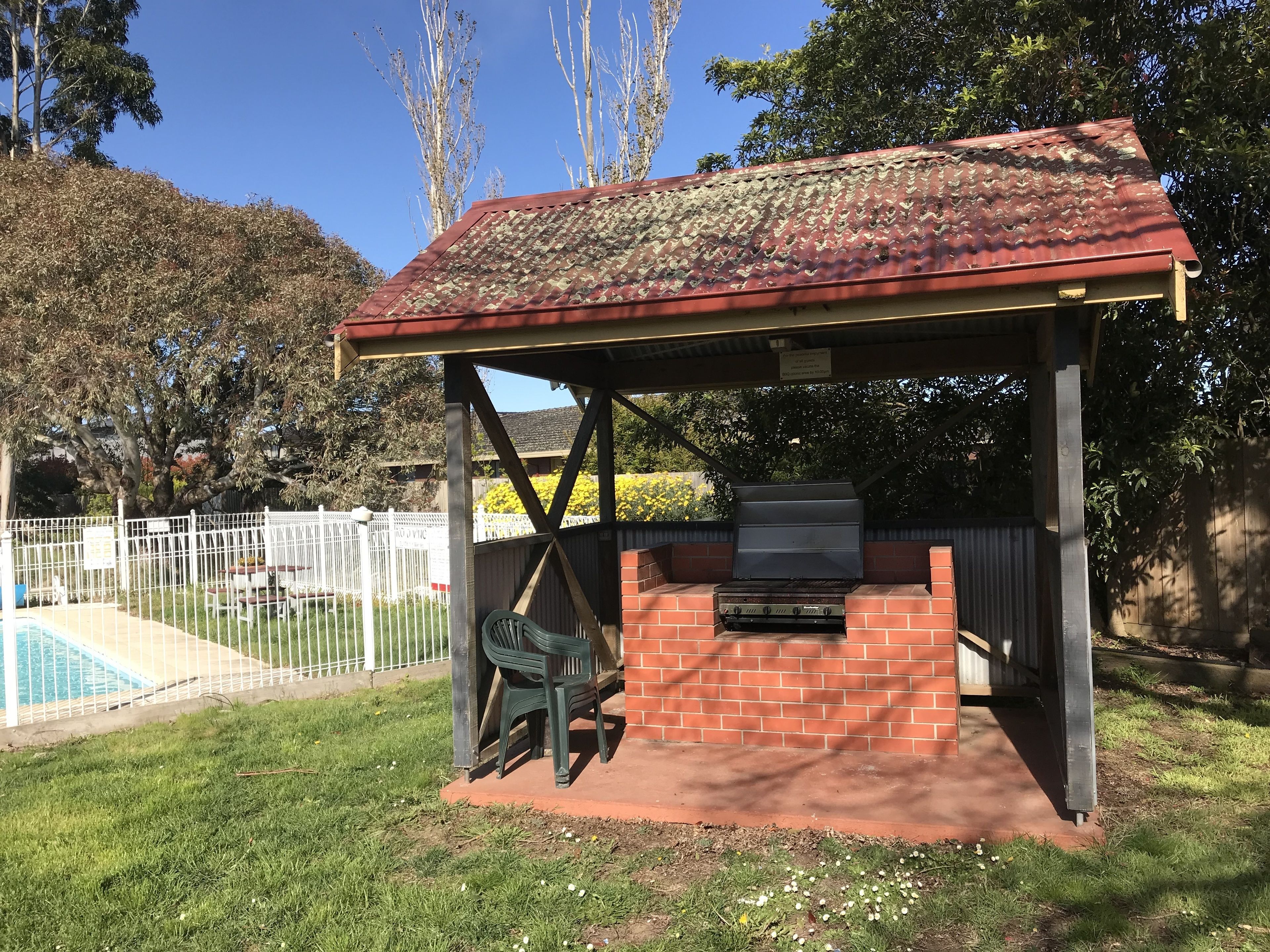 An outdoor barbecue area with a brick grill, a covered seating area, and a pool in the background.