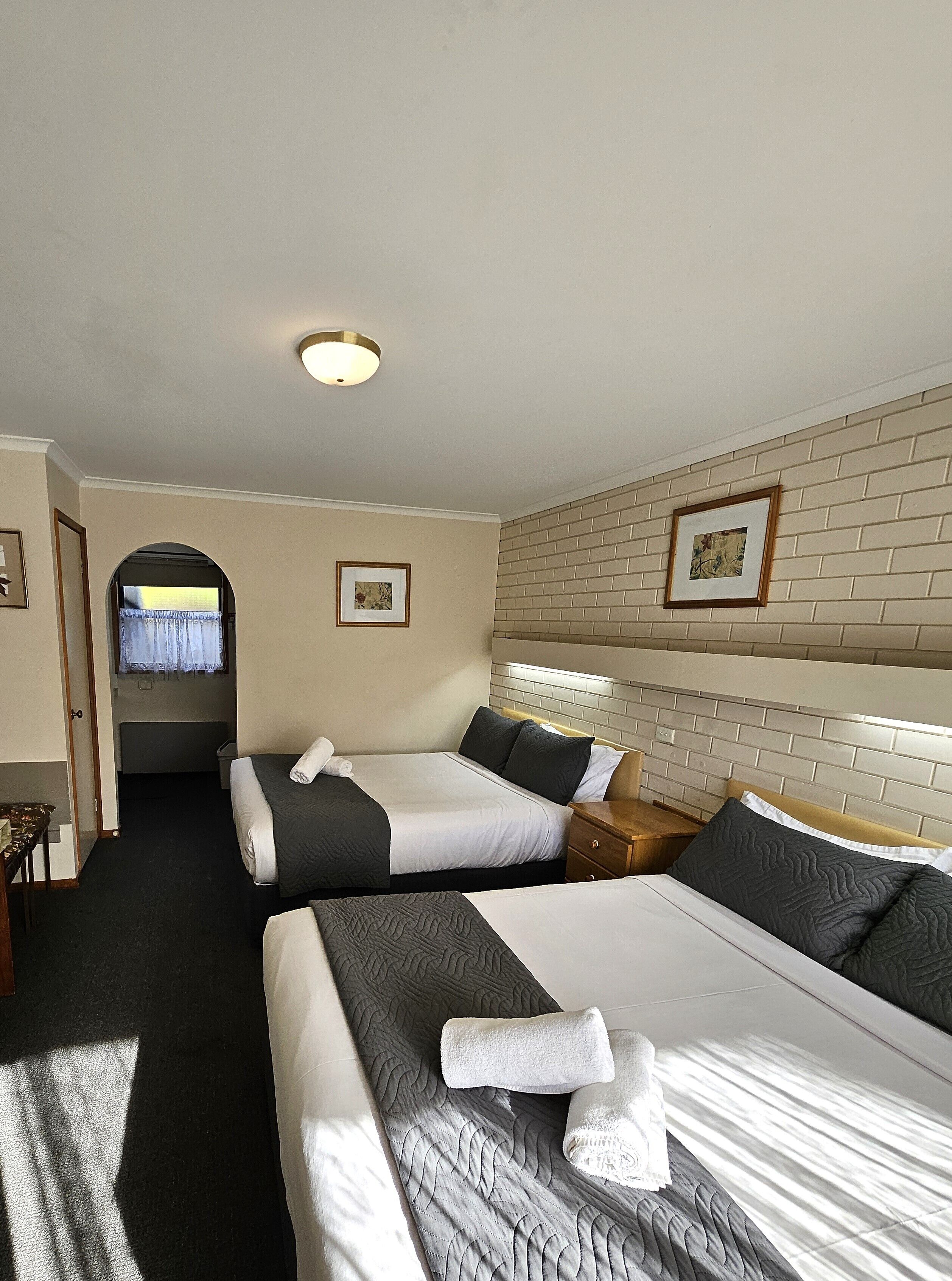 Une chambre d’hôtel avec deux lits, une tête de lit décorée d’un motif en briques, un tableau encadré au mur et une porte donnant sur une autre pièce.