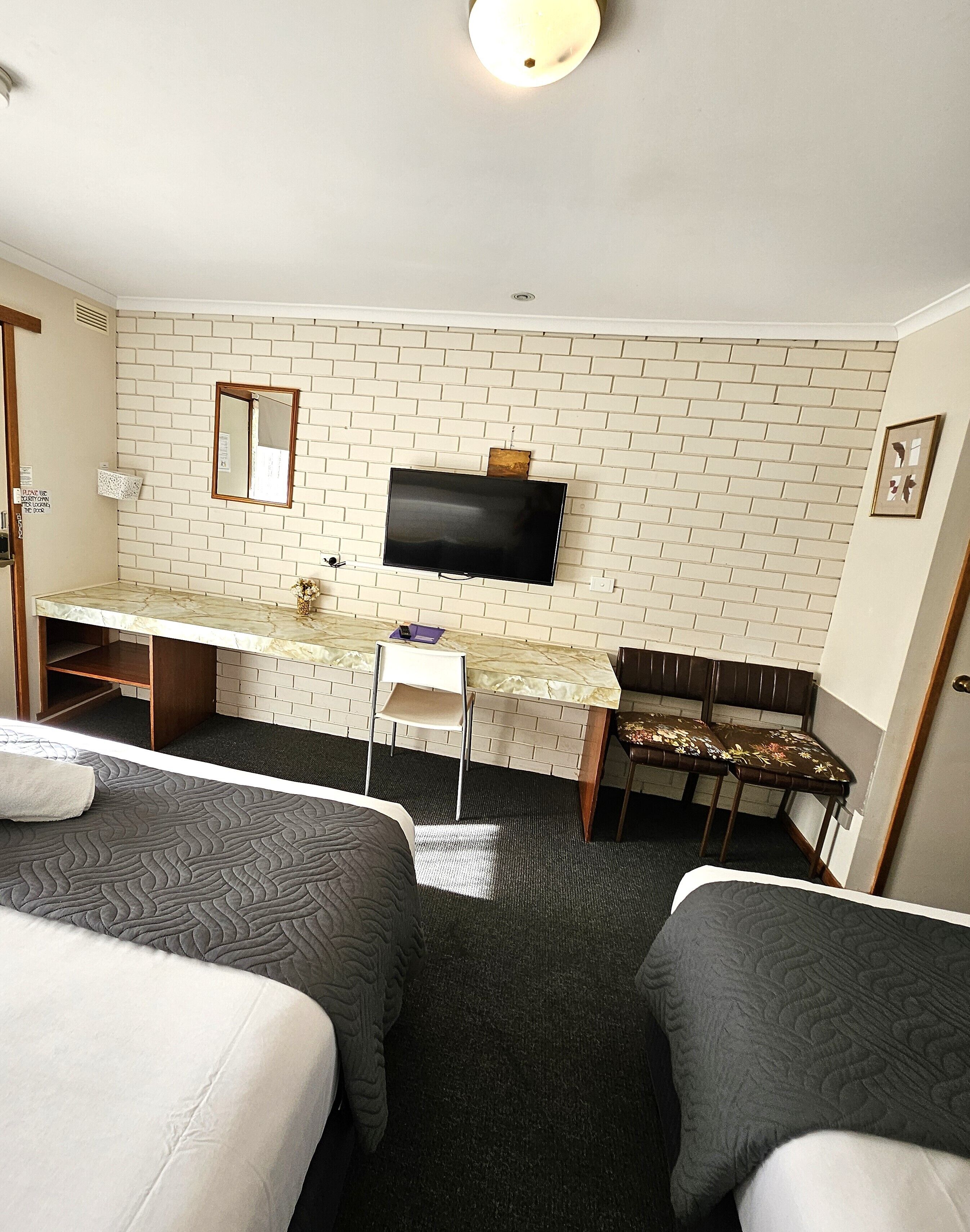 Habitación de hotel con dos camas, un escritorio con silla, un televisor montado en una pared con diseño de ladrillos y una mesita con otra silla.