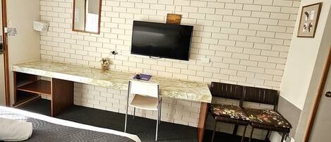 Habitación de hotel con dos camas, un escritorio con silla, un televisor montado en una pared con diseño de ladrillos y una mesita con otra silla.