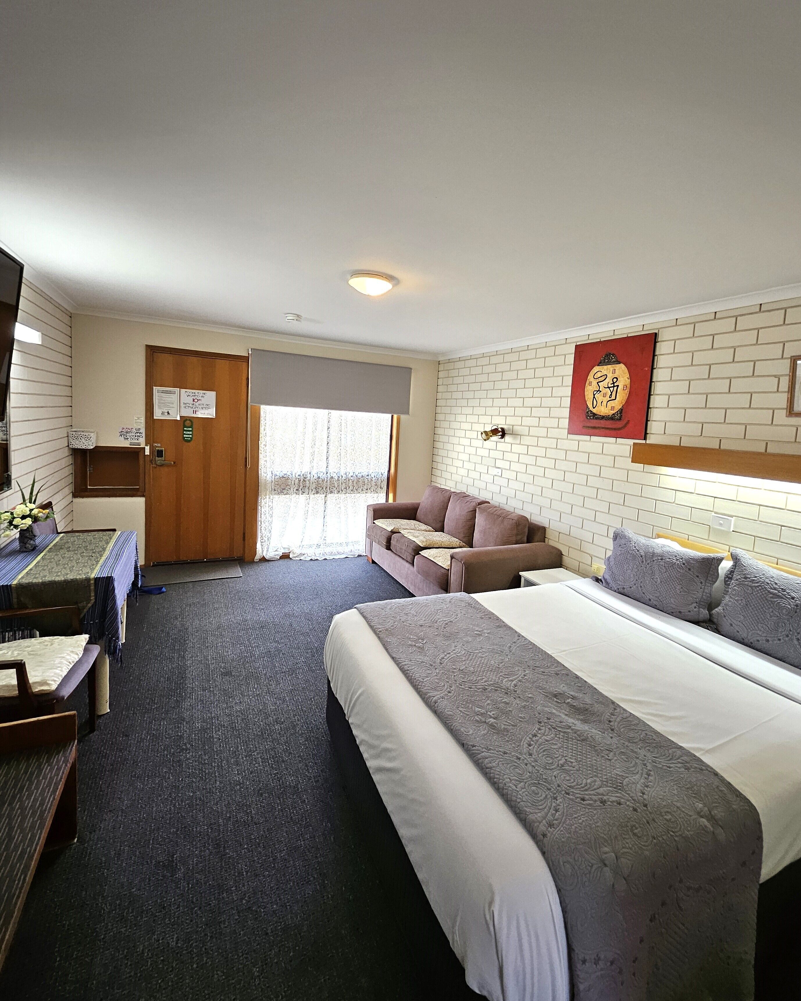 Habitación de hotel con una cama grande, un sofá, un escritorio y una silla. Se ve una ventana con cortinas, una puerta y una pared con una placa decorativa.