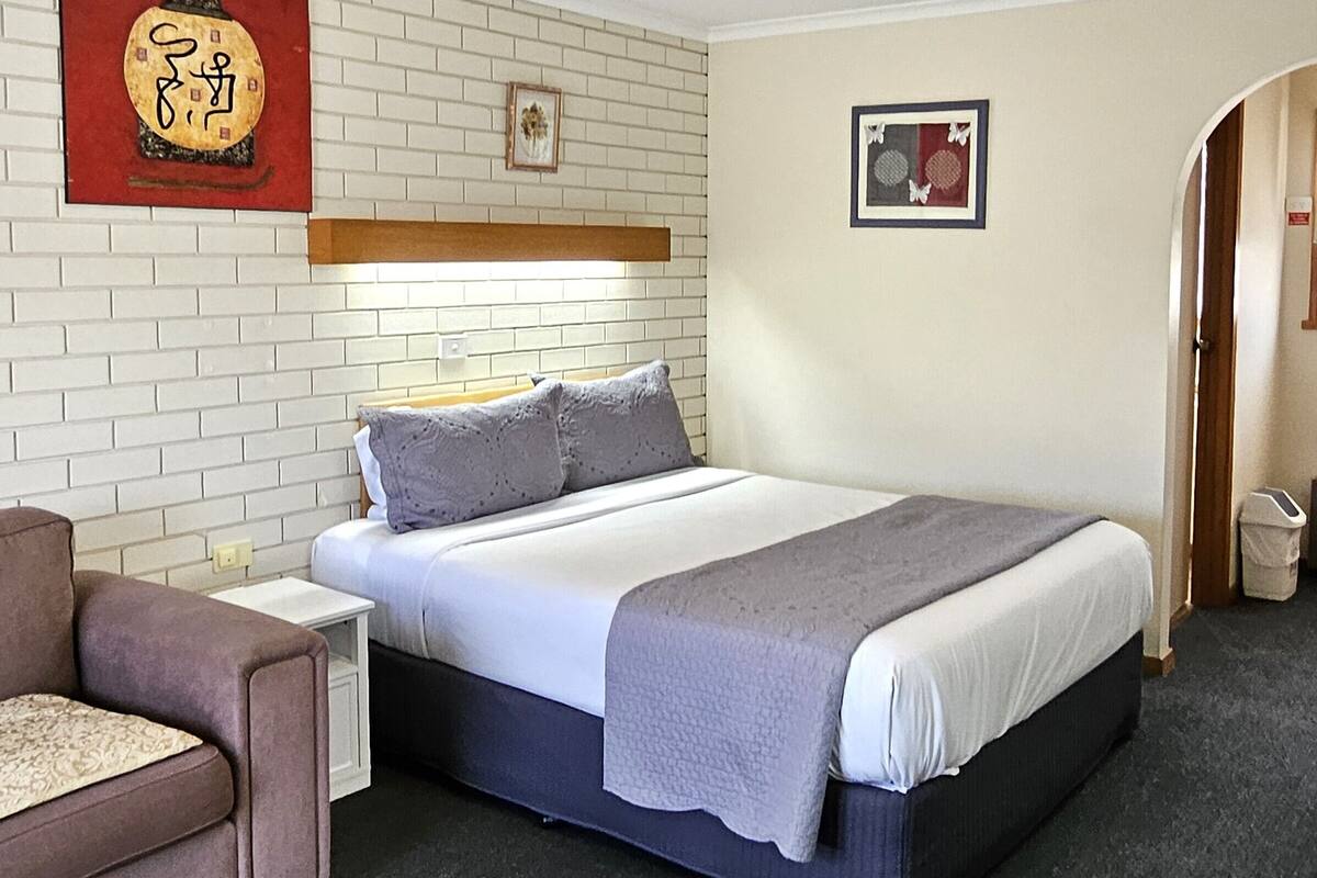 A bedroom with a bed, a nightstand, a chair, and a wall with a picture and a red plaque.