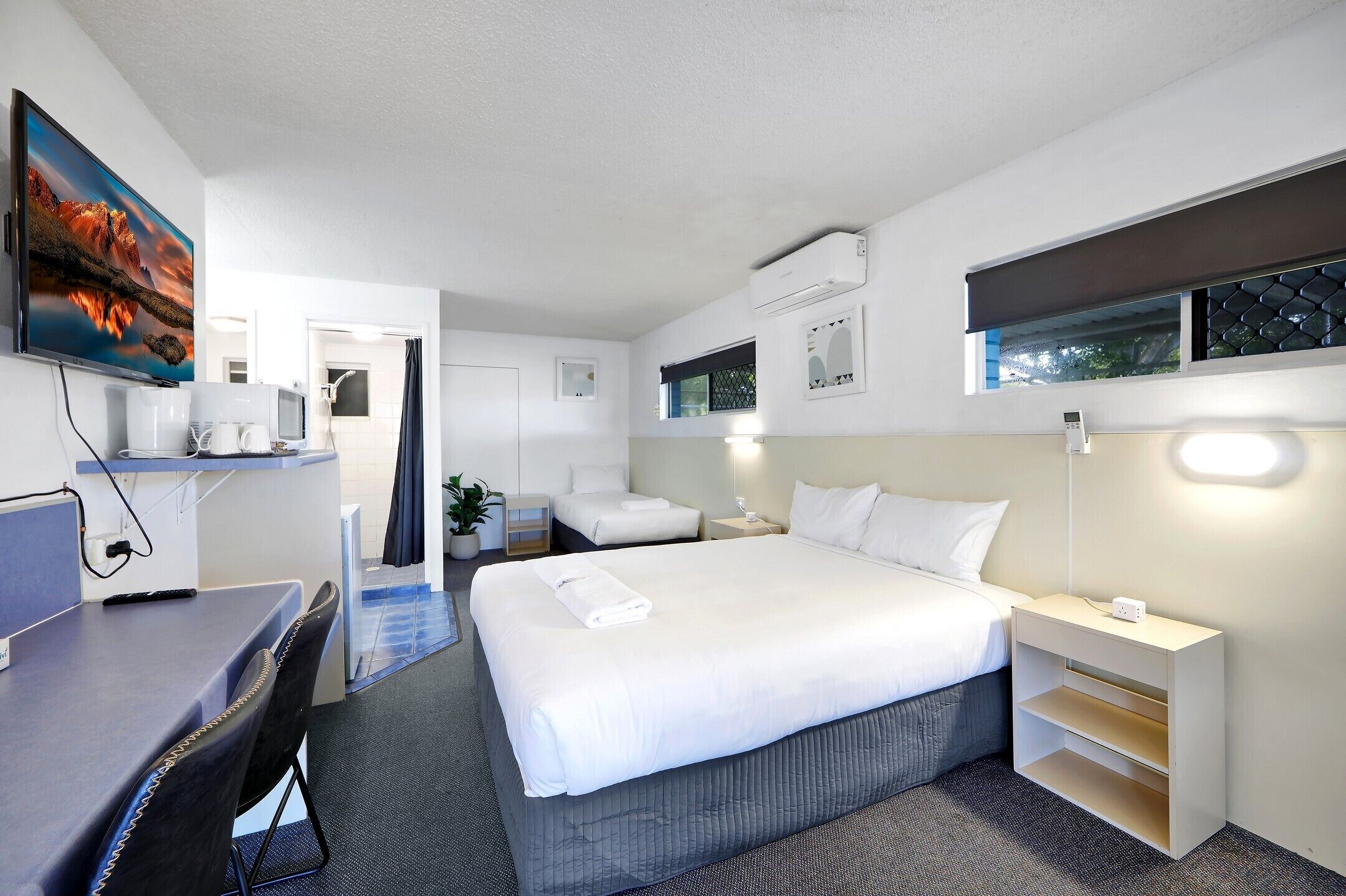 A modern hotel room with a large bed, a desk, a chair, a small table, a TV, and a window with blinds.
