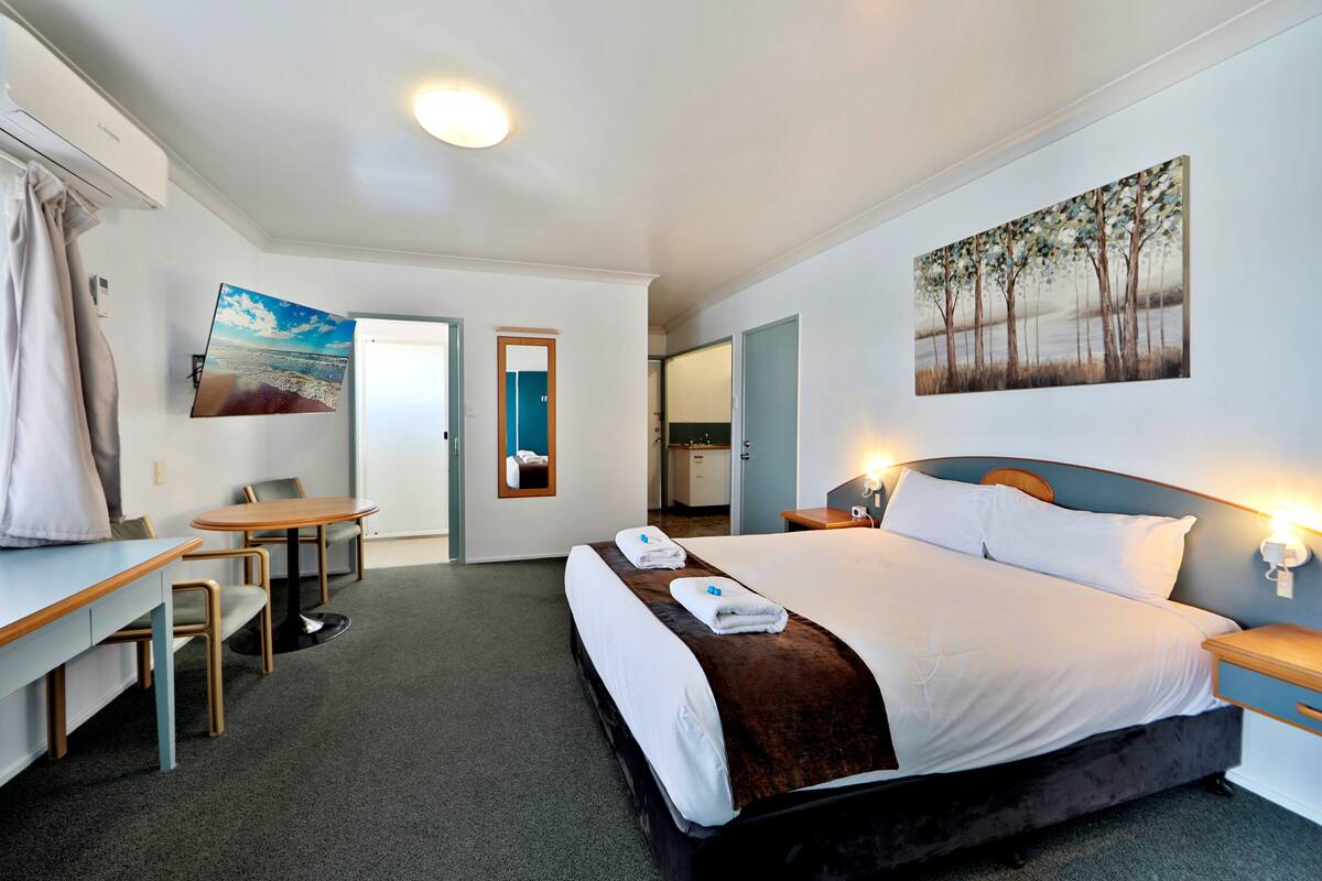 A hotel room with a bed, a desk, a chair, a small table, and a painting on the wall.