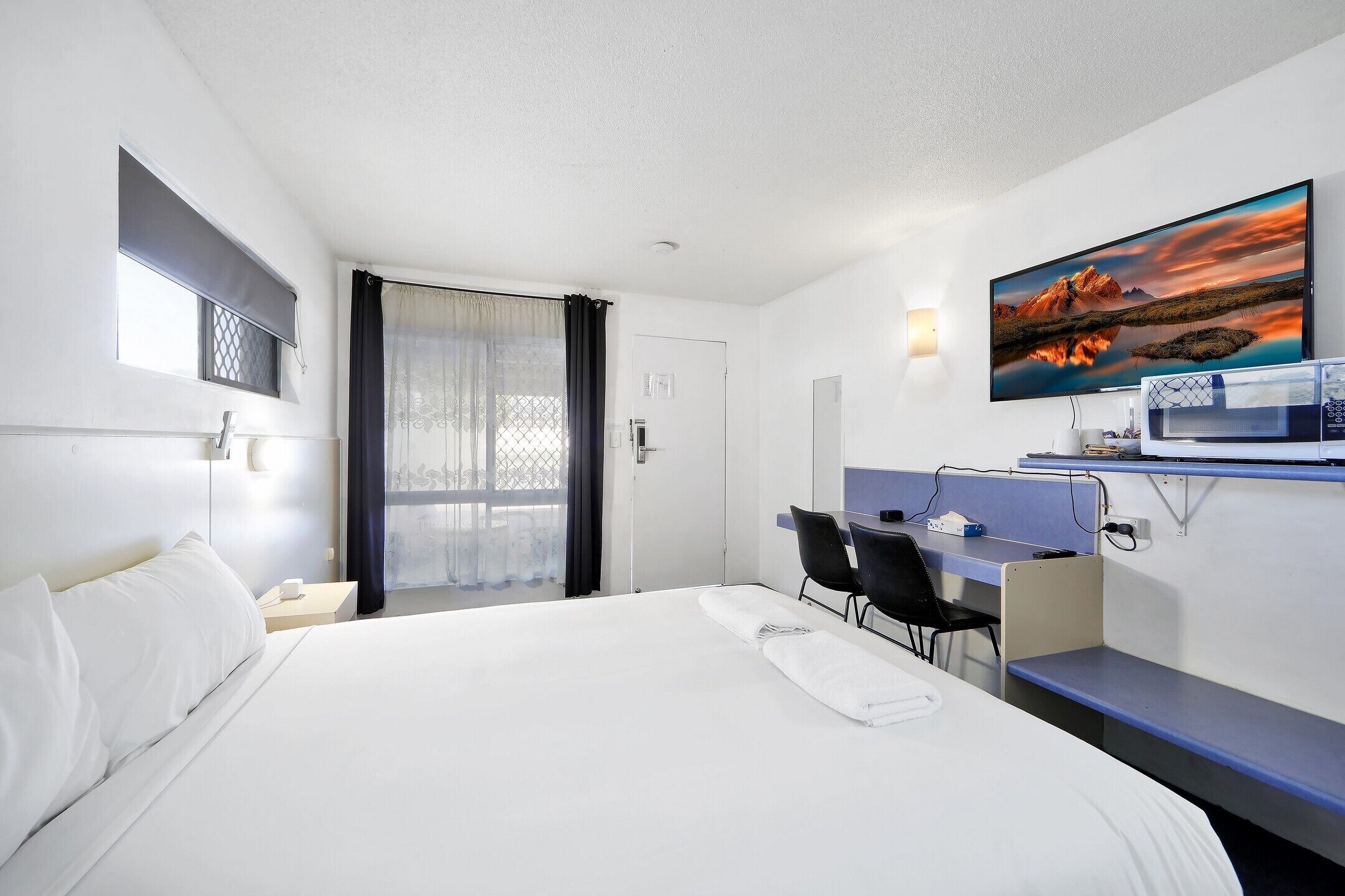 Habitación de hotel con cama, escritorio, silla, televisor y ventana con cortinas.