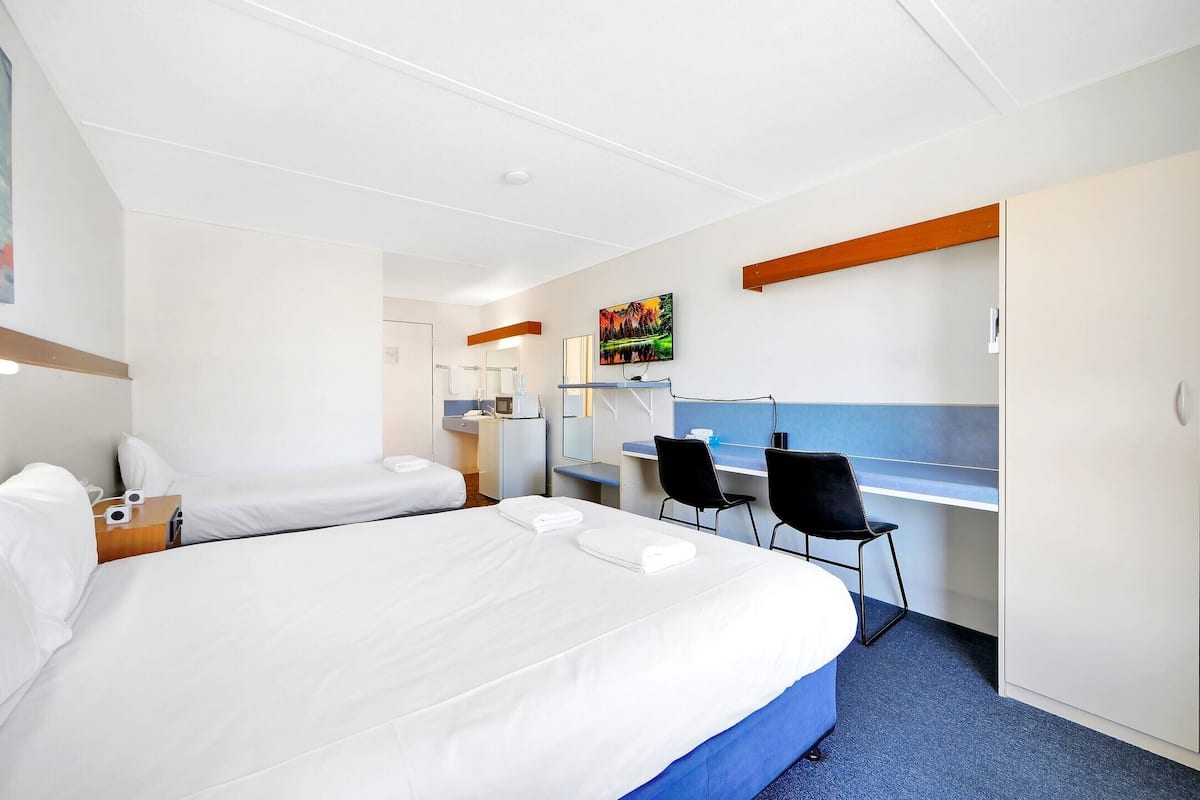 A hotel room with a large bed, a desk, two chairs, a wardrobe, and a painting on the wall.