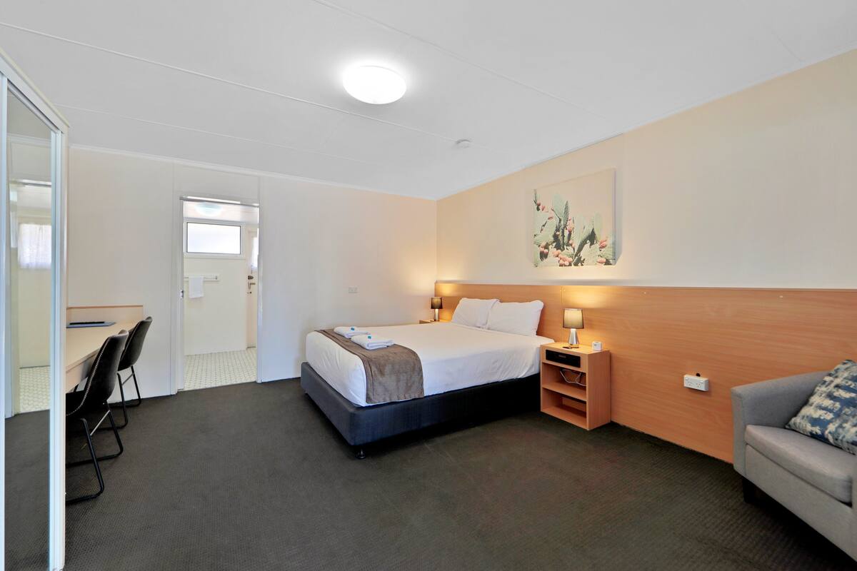 A hotel room with a bed, bedside tables, a desk, a chair, a sofa, and a painting on the wall.
