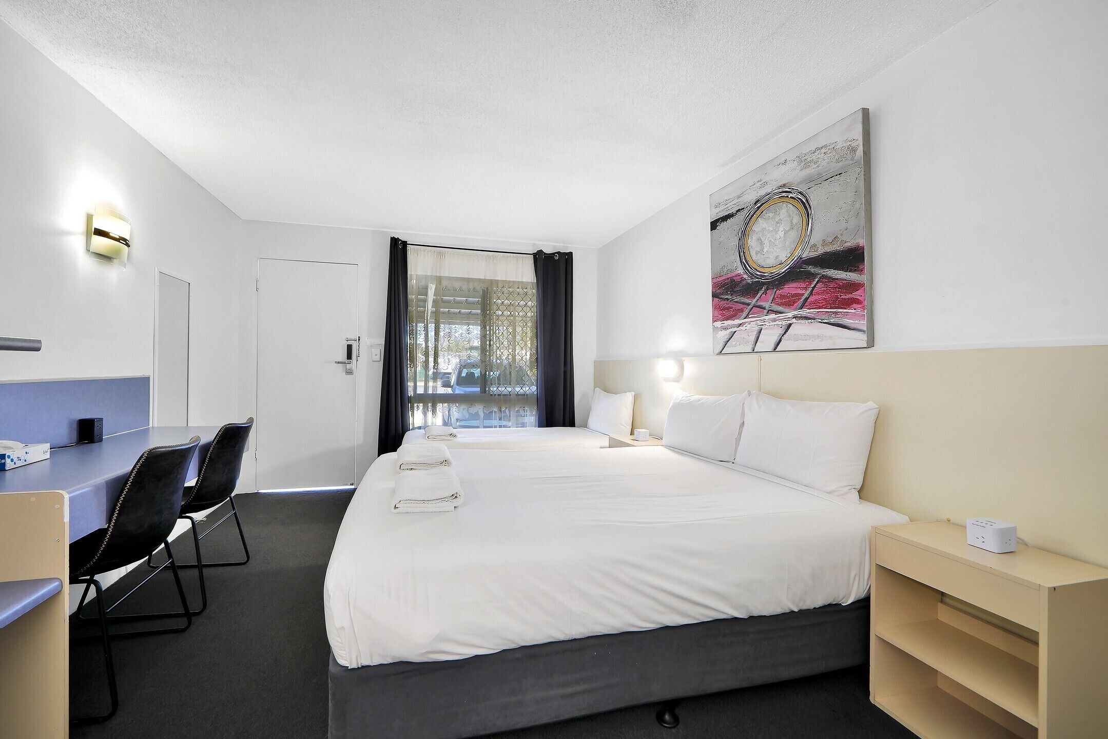 A hotel room with two beds, a desk with chairs, a lamp, and a painting on the wall.