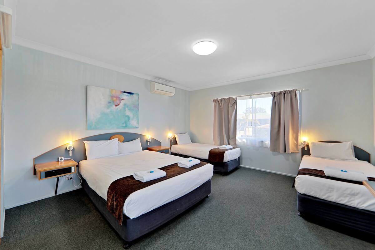 A hotel room with three beds, a painting on the wall, and a window with curtains.