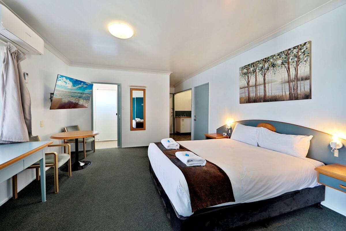 A hotel room with a bed, a desk, a chair, a painting, and a bathroom visible through an open door.