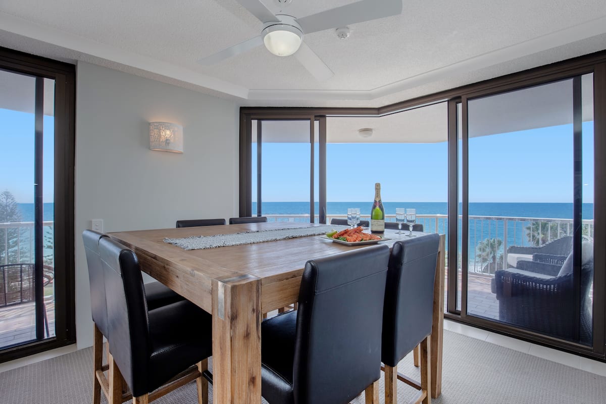 Een eethoek met een houten tafel, zwarte stoelen, een fles wijn en een glas op tafel. Door grote ramen is er uitzicht op de oceaan.