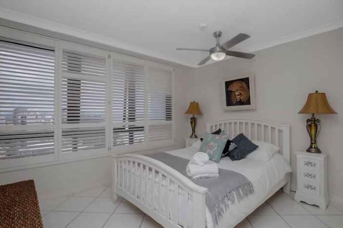 A bedroom with a bed, bedside lamps, a ceiling fan, and a window with blinds.