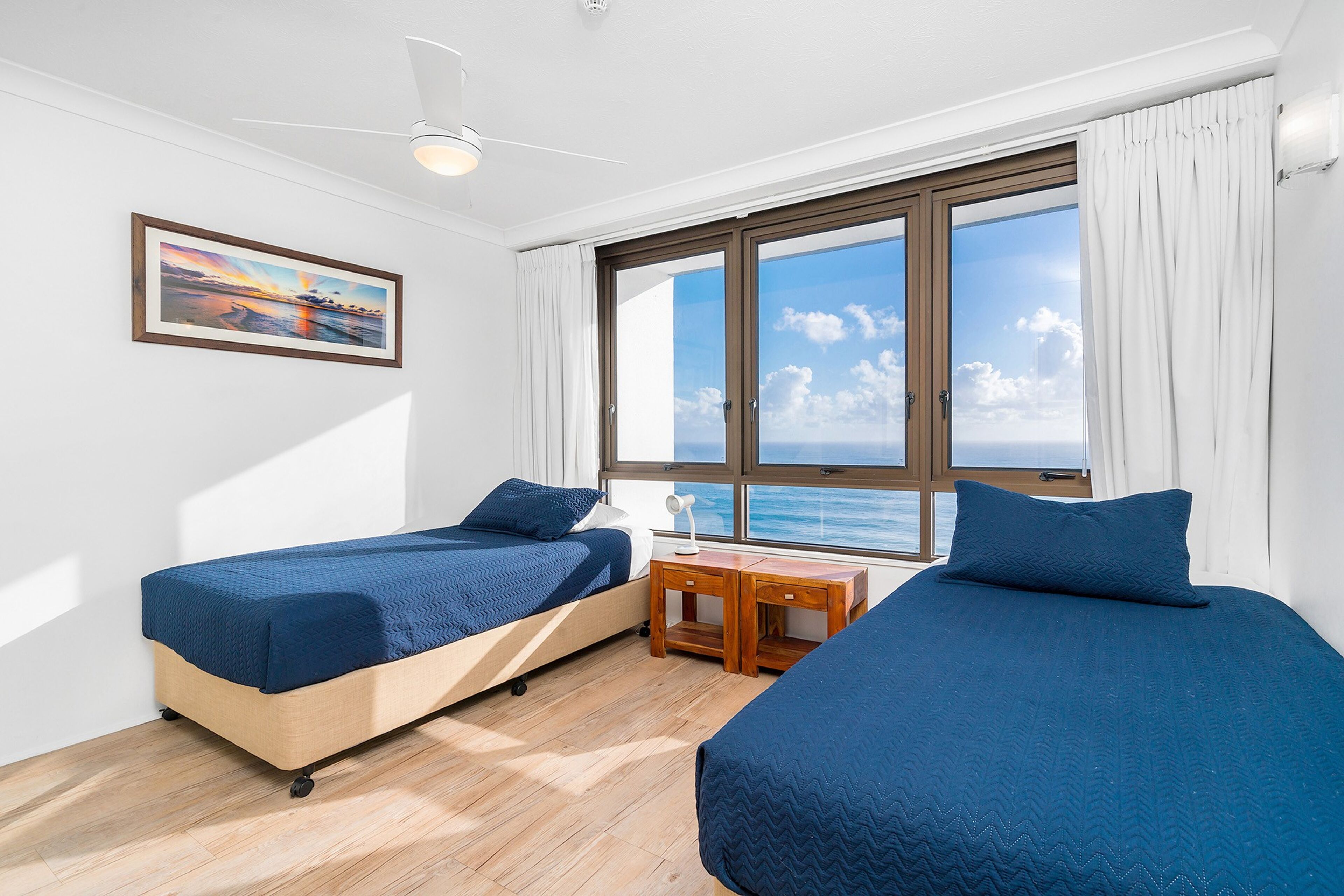 A room with two beds, a wooden nightstand, a framed picture, and a large window with a view of the sea.