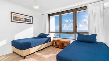 A room with two beds, a wooden nightstand, a framed picture, and a large window with a view of the sea.