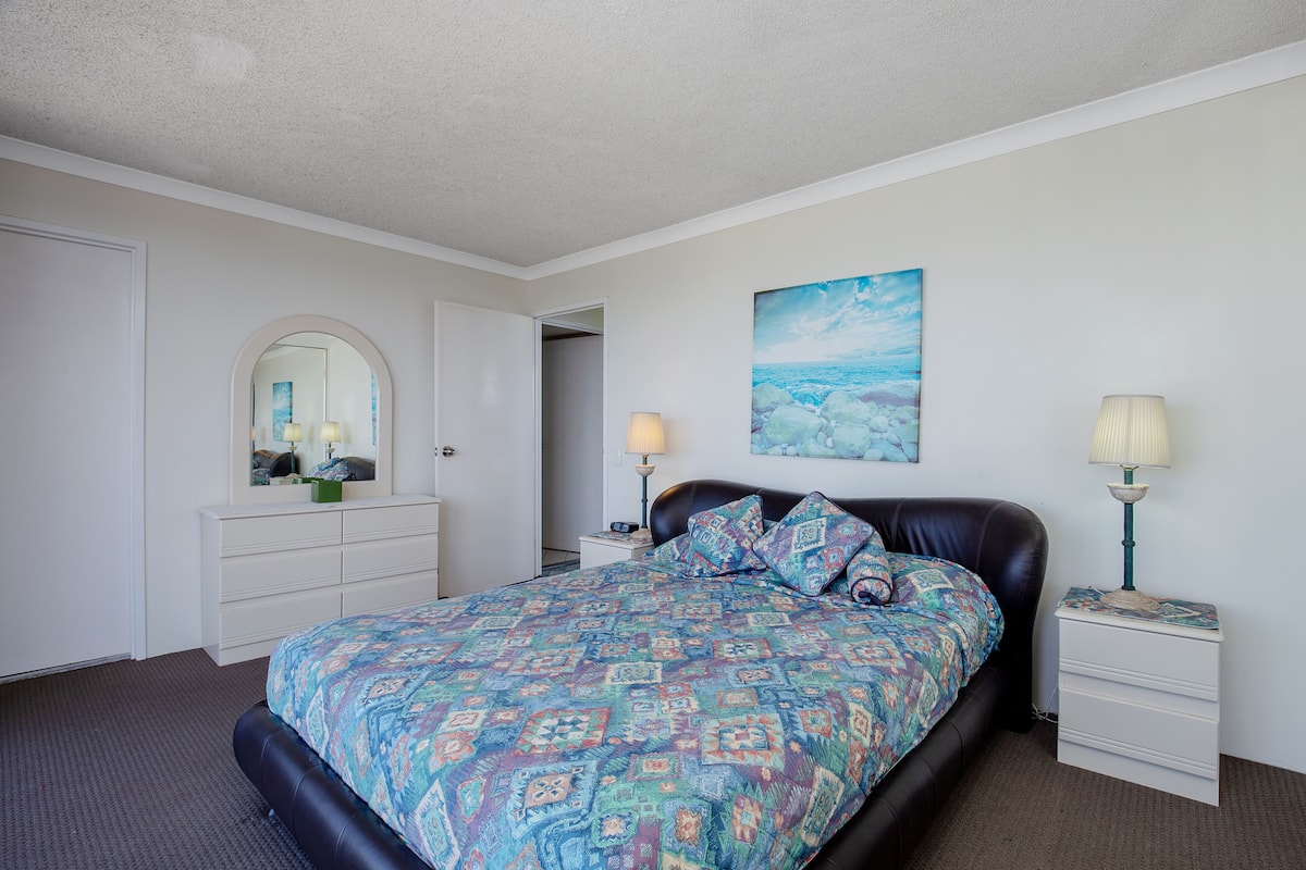 Een slaapkamer met een bed met een geborduurd sprei, een grote spiegel, een nachtkastje met een lamp en een schilderij aan de muur.