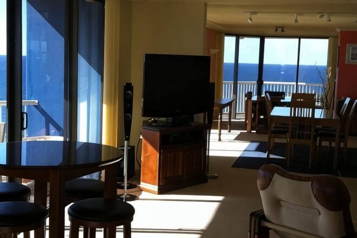 Een kamer met een groot raam, een televisie op een houten standaard, een eettafel met stoelen en een balkon met een hekwerk.